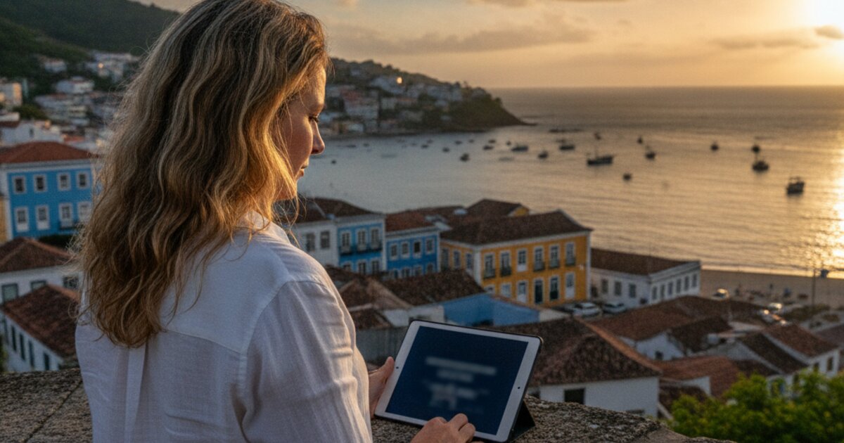 Mulher branca de cerca de 40 anos, de costas, olhando um tablet em um mirante sobre a baía com a cidade histórica ao fundo